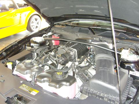 2007 Ford Mustang - Engine bay of a black car with a supercharger, a yellow car is visible in the background.