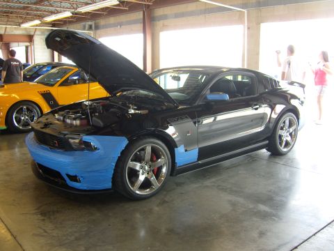2007 Ford Mustang - A black Ford Mustang is parked with its hood up, showing blue tape on the front bumper. Another yellow Mustang is visible behind it.
