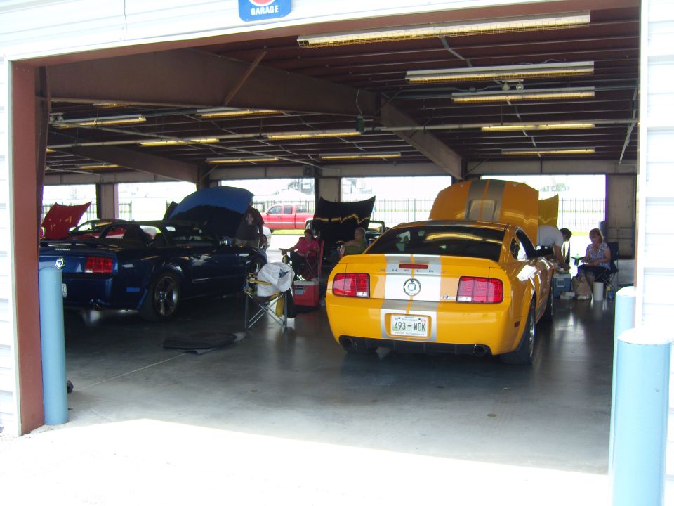 Yellow 2007 Ford Mustang GT with silver stripes parked in garage next to a blue Mustang