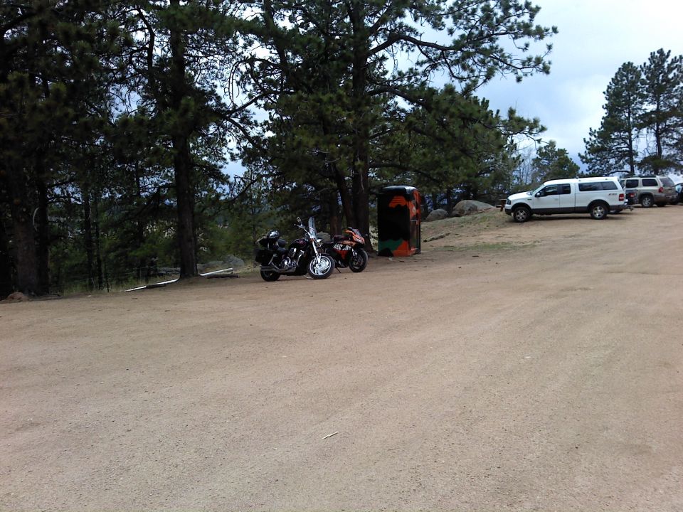 Two motorcycles parked near trees, with SUVs in the background on a dirt lot