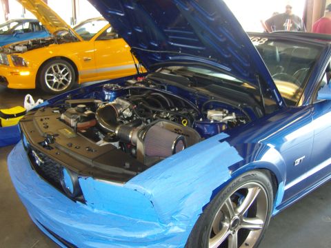 2007 Ford Mustang - A blue Ford Mustang GT's engine bay is visible, with the front end covered in blue tape, while a yellow Mustang sits behind it.