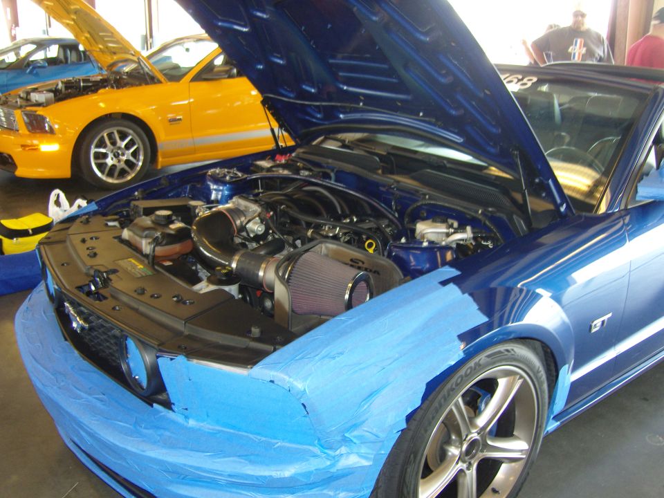 Blue Ford Mustang GT with hood open, engine exposed, front bumper taped off. Yellow Mustang in background