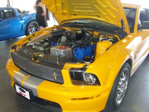 2007 Ford Mustang - A yellow Ford Mustang is parked with its hood open, revealing the engine bay. A blue Chevrolet Corvette is visible in the background.