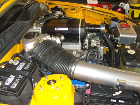 2007 Ford Mustang - The engine bay of a yellow 2007 Ford Mustang shows a prominent supercharger and custom intake system.