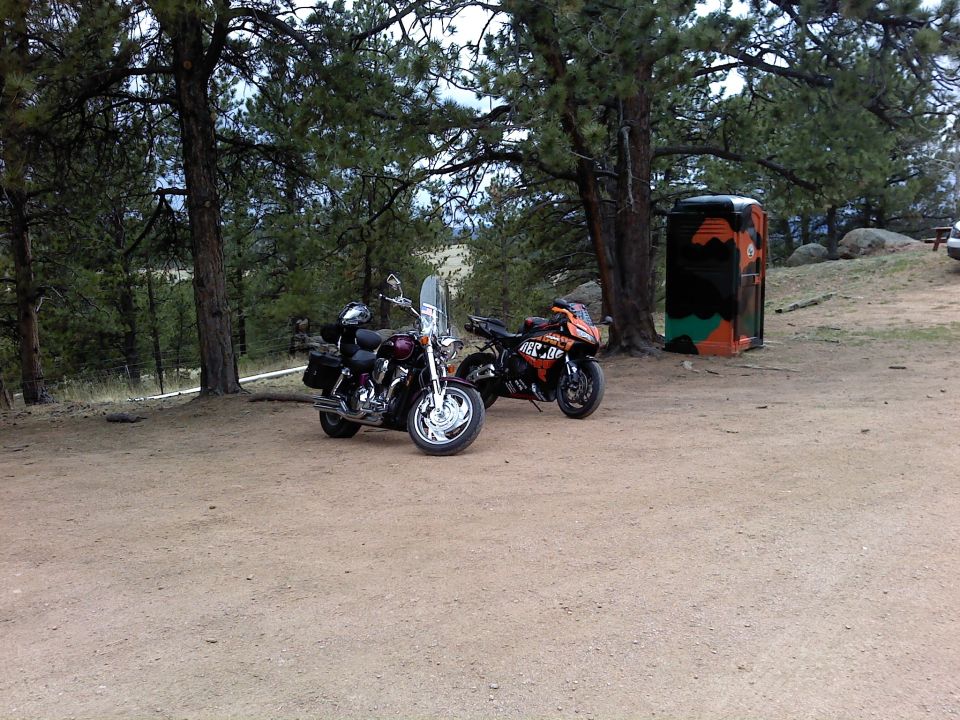 Two motorcycles parked on a dirt lot near trees and a portable toilet