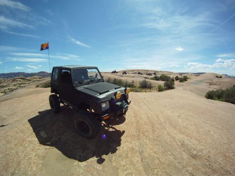 1987 Suzuki Samurai - Wide-angle view of a lifted black Suzuki Samurai hardtop parked on open slickrock under a blue sky, flying an Arizona state flag from a rea…