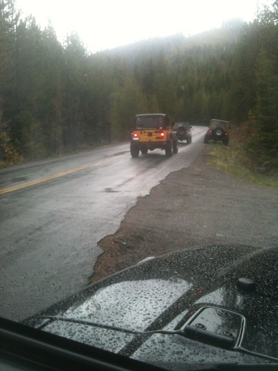 Several Jeeps driving on a wet, tree-lined road. The lead Jeep is yellow