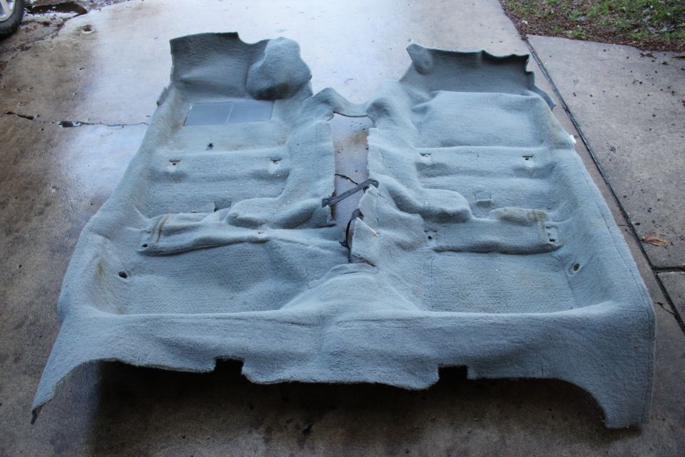 Gray molded automotive floor carpet removed from a car, laid out on wet concrete driveway