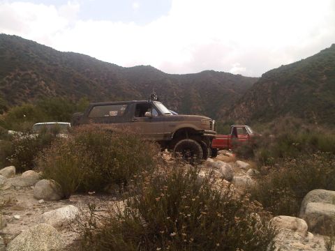 1986 Ford Bronco - A lifted brown Ford Bronco navigates a rocky mountain path, with another truck visible in the background.