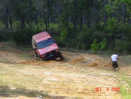 1991 Ford Bronco - A red SUV is stuck in the mud on a dirt trail with a person running in the background.