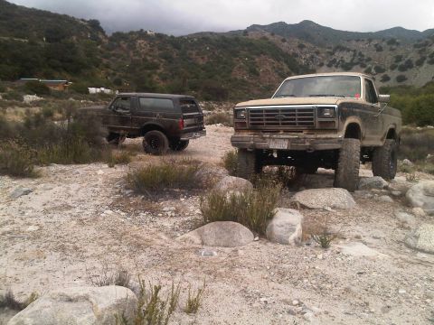 1986 Ford Bronco - Two Ford trucks, a Bronco and an F-Series, are parked off-road in a dusty, mountainous area.