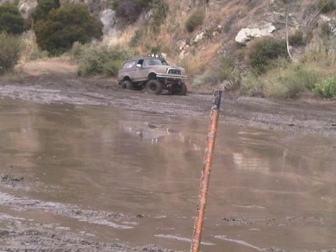 1986 Ford Bronco - This lifted Ford Bronco is tackling a challenging muddy terrain, showing its off-road capabilities.