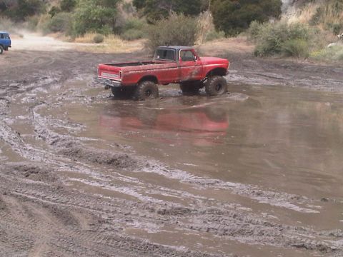 1986 Ford Bronco - A red pickup truck navigates a muddy off-road course, with another vehicle visible in the distance.
