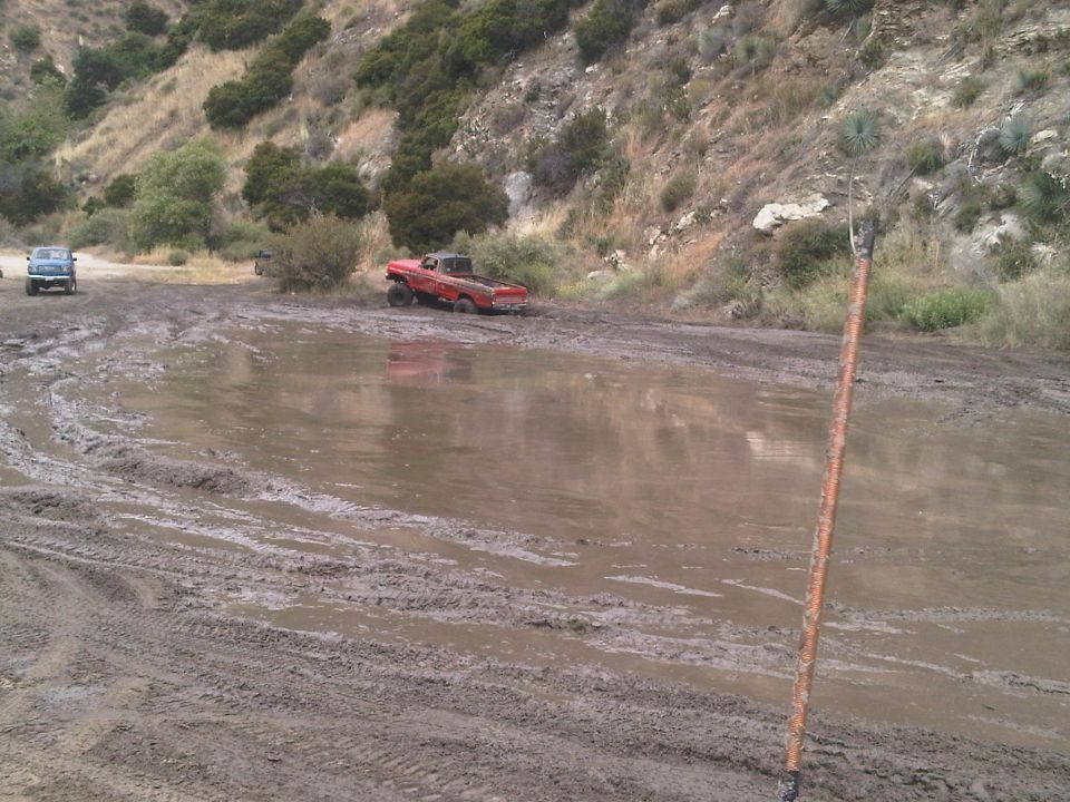 Red pickup truck stuck in a large mud puddle with another vehicle in the background