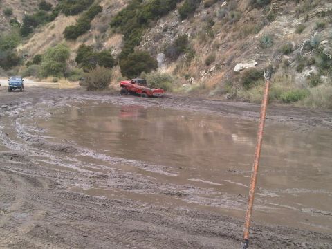 1986 Ford Bronco - A red pickup truck navigates a muddy terrain, with another vehicle in the distance.