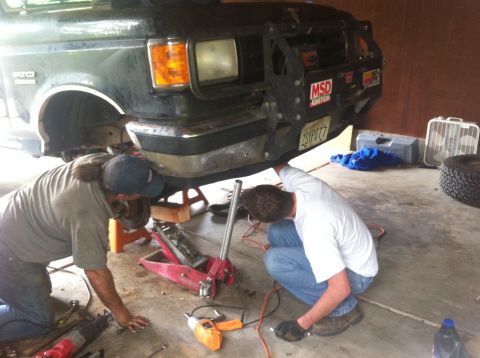 1990 Ford Bronco - Two mechanics work on the front end of a dark green Ford Bronco, which appears to be from the late 1980s.