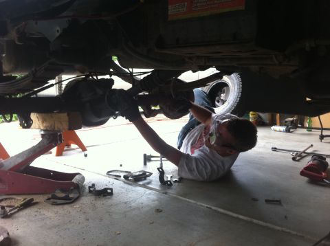 1990 Ford Bronco - A person is working on the underside of a vehicle that is supported by jack stands and a floor jack.