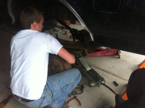 1990 Ford Bronco - A mechanic works on the rear suspension of a black SUV, with tools and jacks visible on the ground.