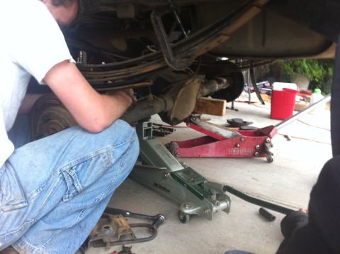 1990 Ford Bronco - Mechanic kneeling beneath a raised vehicle, working on the undercarriage with a red floor jack and an ACDelco jack visible on a concrete dr…