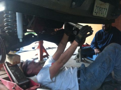 1990 Ford Bronco - Mechanics work on the suspension of a vehicle that is raised on jack stands and a floor jack.