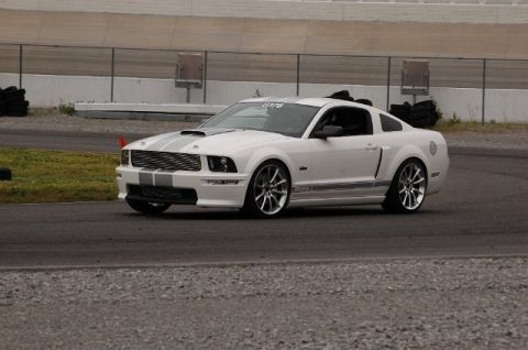 2007 Ford Mustang - White 2007 Ford Mustang with gray racing stripes on a track during an event.