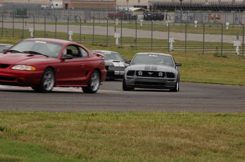 2007 Ford Mustang - A gray Ford Mustang with racing stripes drives on a track, with a red Mustang in the foreground.