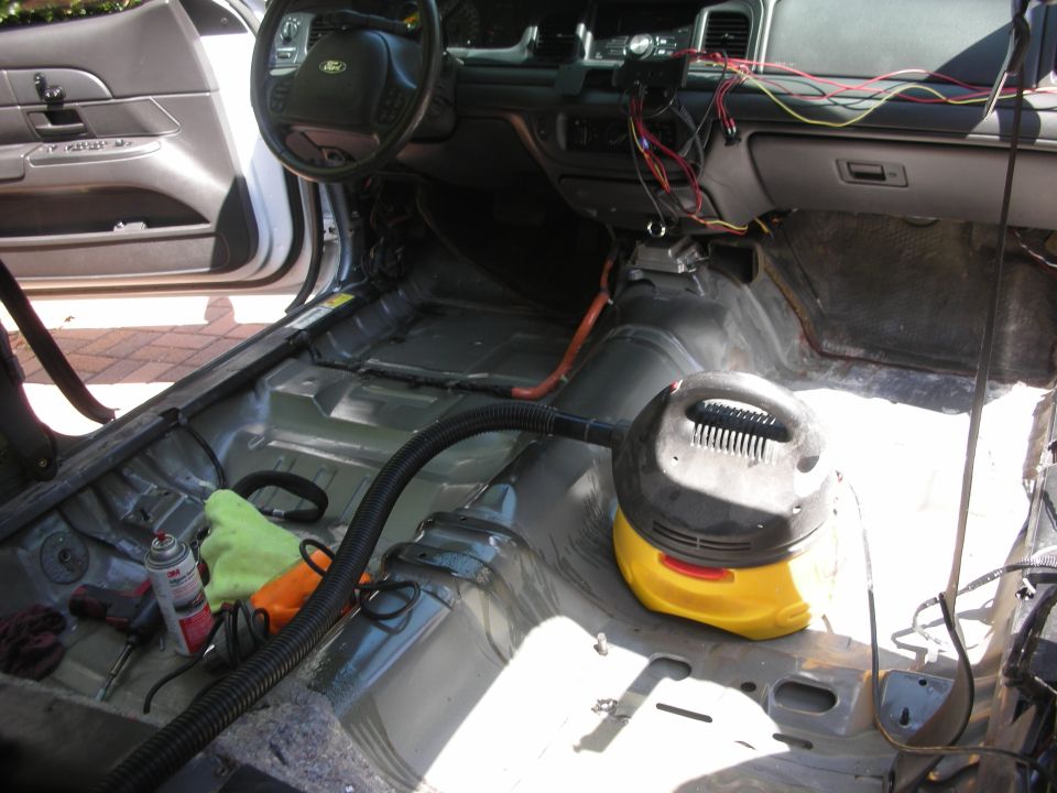 Interior of a 2004 Ford Crown Victoria with seats removed, showing exposed metal floor and wiring, with a vacuum cleaner and tools