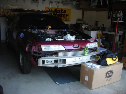 1992 Ford Crown Victoria - A maroon 1992 Ford Crown Victoria is partially disassembled in a garage, with its front bumper removed and engine components visible.