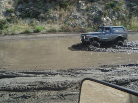 1986 Ford Bronco - A lifted Ford Bronco navigates a muddy terrain, kicking up water and dirt.