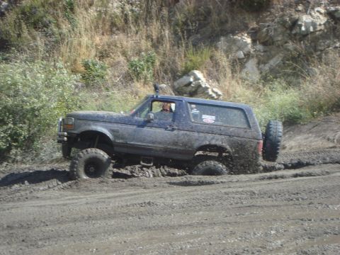 1986 Ford Bronco - A blue Ford Bronco navigates a muddy off-road trail, with its tires deeply embedded in the mire.