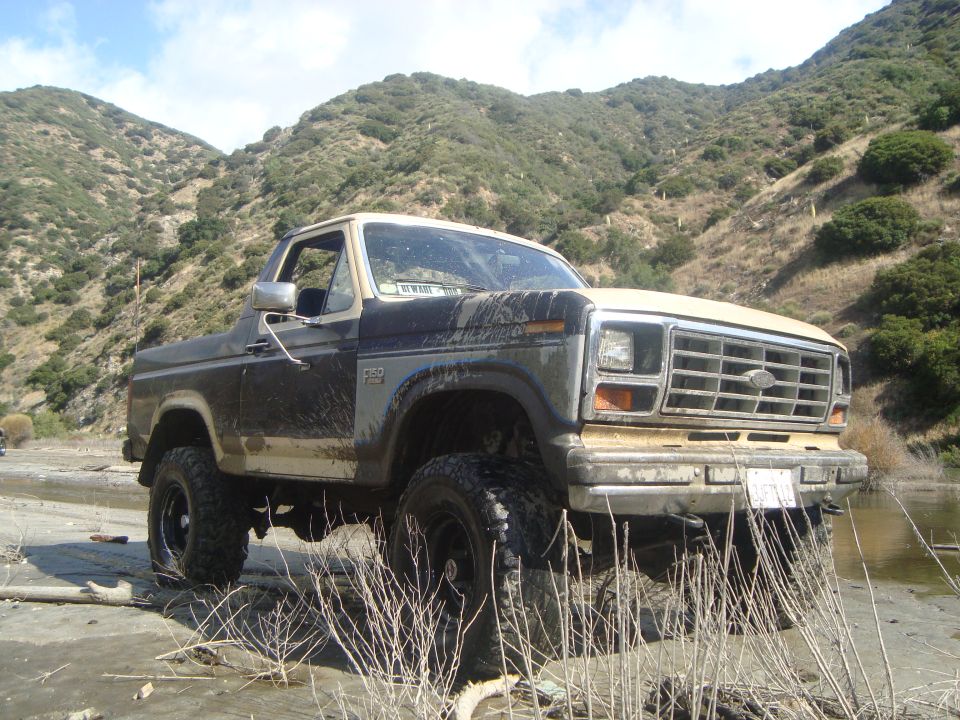 Ford Bronco parked in dry riverbed with muddy tires and body, hills in background