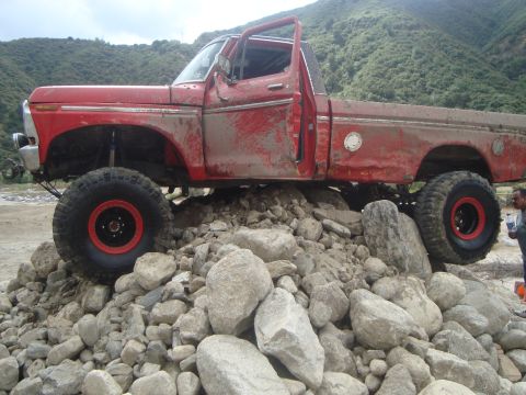 1986 Ford Bronco - This lifted red pickup truck is navigating a rocky terrain, with its doors ajar and covered in mud.