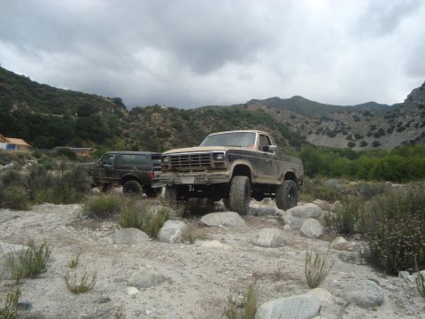 1986 Ford Bronco - Two off-road vehicles are parked in a rocky, mountainous area under a cloudy sky.