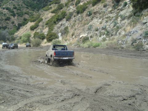 1986 Ford Bronco - Off-road vehicles navigate a muddy track in a hilly, arid landscape.