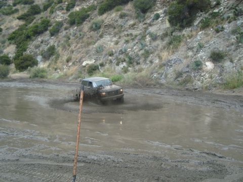 1986 Ford Bronco - A dark SUV navigates a deep mud puddle on a rough, unpaved road.