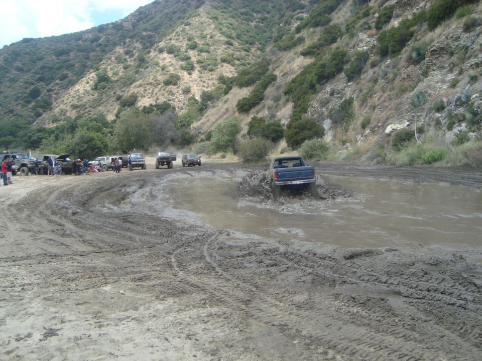 Blue pickup truck splashing through mud, other vehicles gathered on dirt track