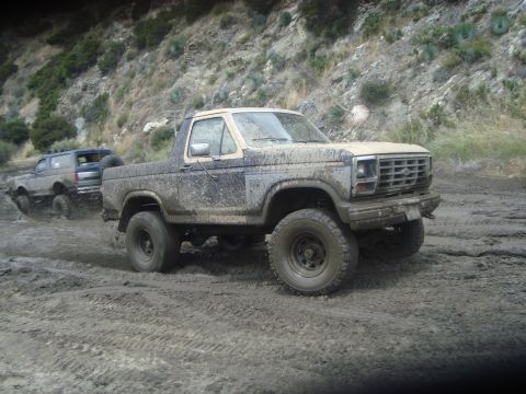 1986 Ford Bronco - Two muddy Ford Broncos navigate a dirt track on a hillside.