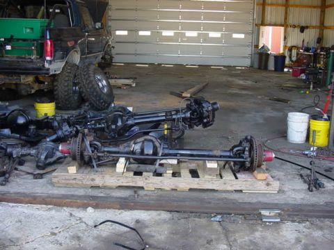 1992 Ford Bronco - Multiple front axles and suspension components are laid out on wooden pallets inside a garage, with a truck visible in the background.