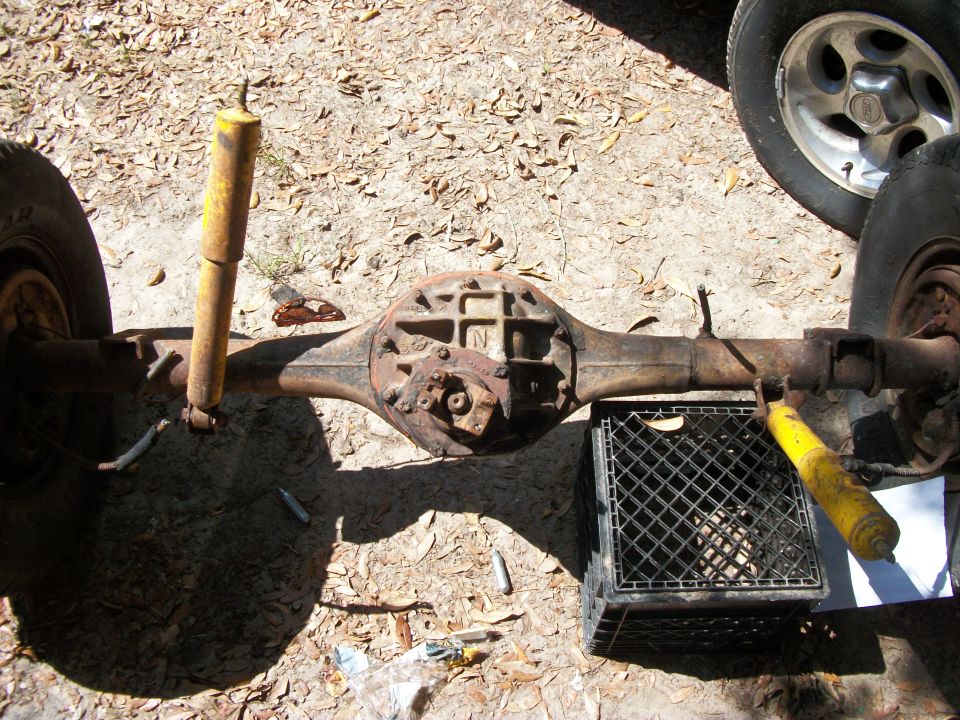 Rusty rear axle housing removed from a truck lying on sandy ground with yellow shocks attached
