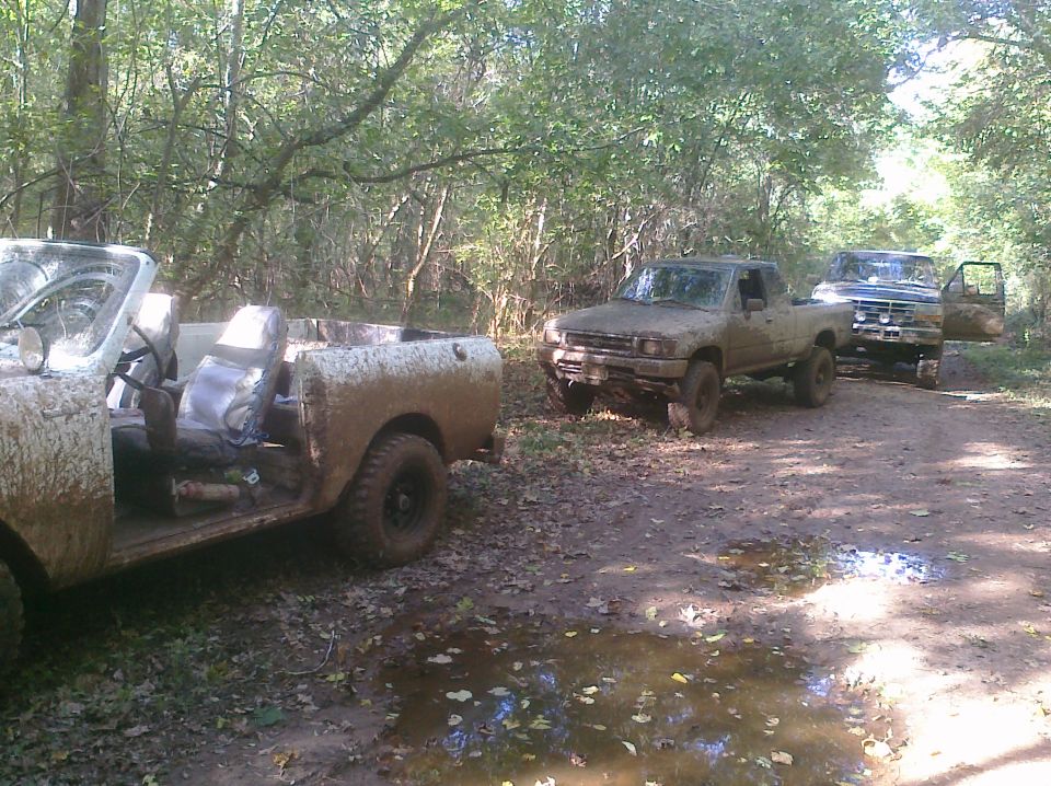 Three muddy off-road vehicles on a dirt path; the closest is a convertible, followed by two pickup trucks