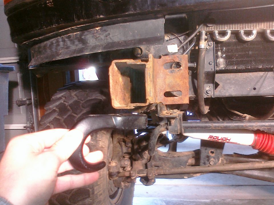 Hand holding black tow hook near rusted front bumper bracket and tire