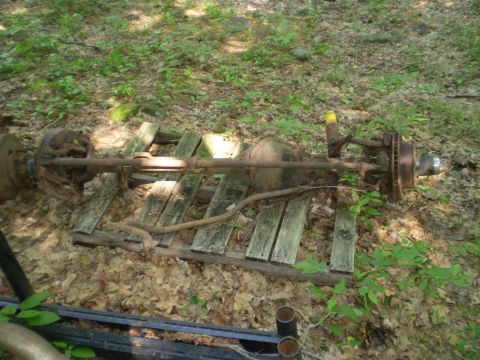 1979 Ford Bronco - Two rusty vehicle axles are resting on weathered wooden planks in a wooded area.