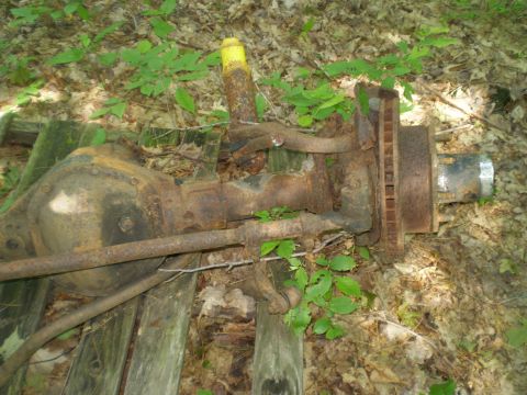 1979 Ford Bronco - This heavily rusted axle assembly, likely from an older vehicle, is partially obscured by foliage and rests on weathered wooden planks.