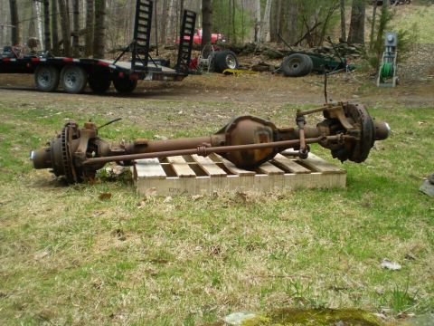 1979 Ford Bronco - A rusty front axle assembly is displayed on a wooden pallet outdoors, with a trailer and trees in the background.