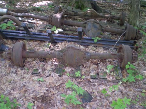 1979 Ford Bronco - Several rusty rear axles are visible, lying on the ground in a wooded area.