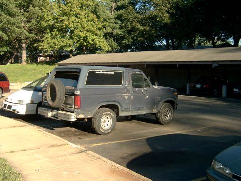 1982 Ford Bronco - A dark blue Ford Bronco is parked in a lot next to a white car, with trees and a covered parking area in the background.
