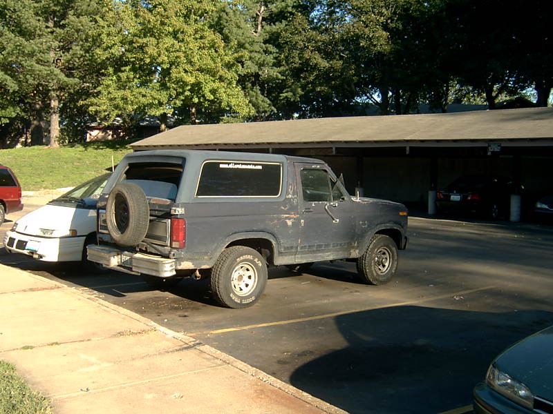 Faded blue Ford Bronco in parking lot, rear glass up, tailgate spare on white wheel