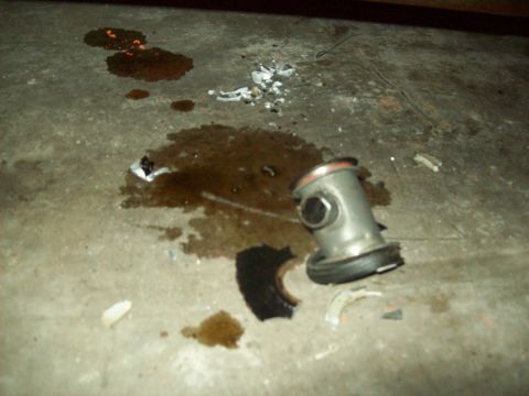 1985 Ford Bronco - A damaged automotive part lies on a concrete floor amidst various spills and debris, suggesting a recent mechanical failure.