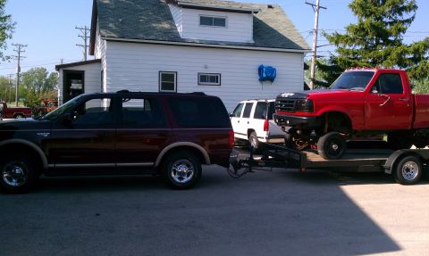 1999 Ford Expedition - A maroon Ford Expedition tows a trailer carrying a lifted red pickup truck, with a white SUV visible behind it.