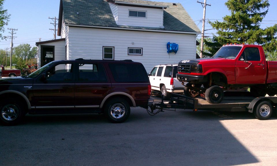 Maroon Ford Expedition towing a trailer with a lifted red pickup truck on it. White SUV behind Expedition